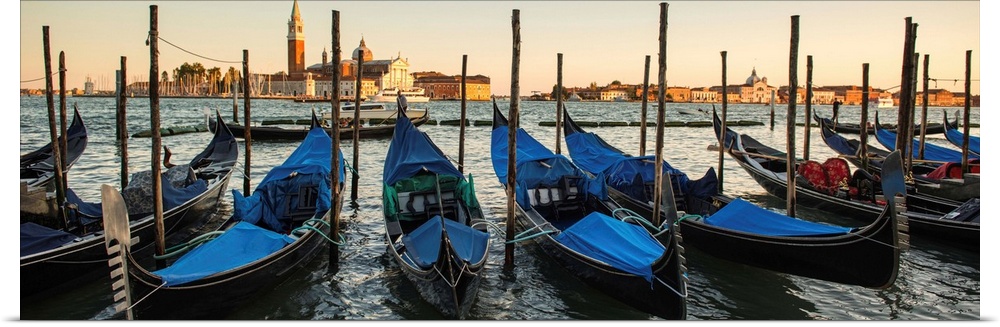 Panoramic photograph of a row of docked gondolas with St. Mark's Square (Piazza San Marco) in the background at golden hour.