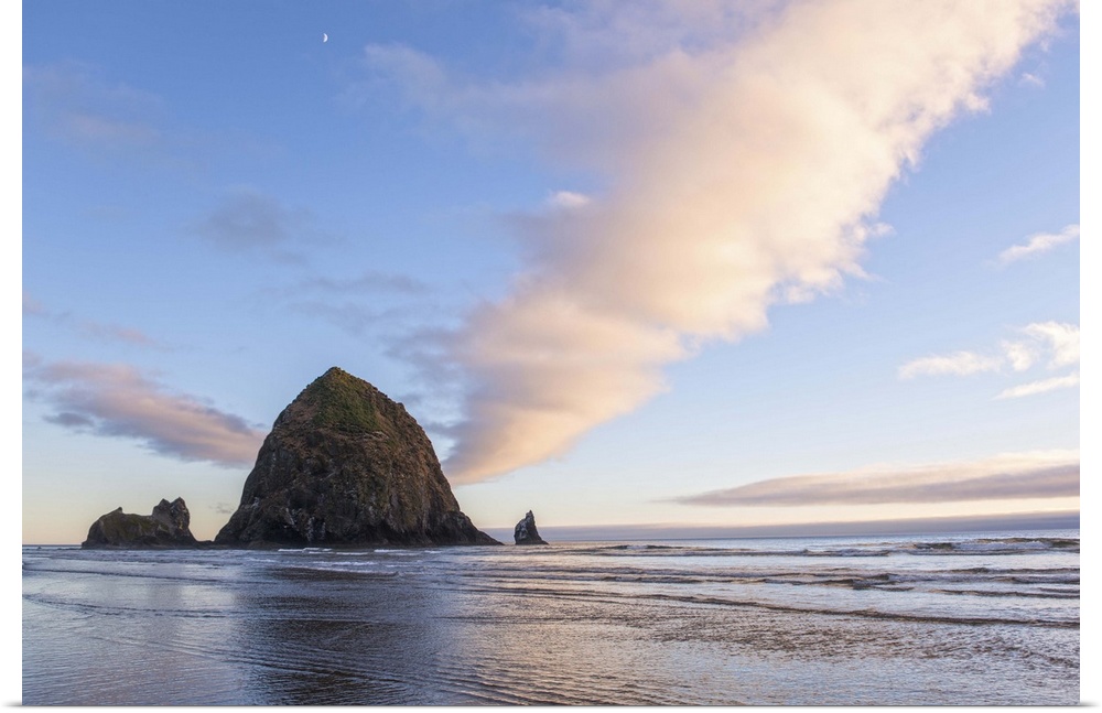 Photograph of Haystack Rock at sunset with rippling waters in the foreground and the moon above.