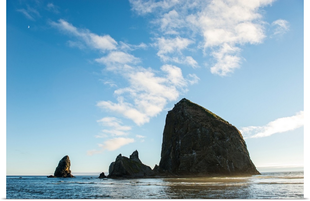 Photograph of Haystack Rock with the golden sun shining on its side and the moon in the top corner.