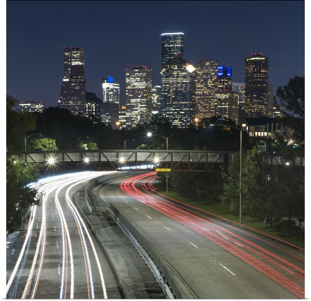 Square photograph of the Houston, TX skyline at night with curving light trails from cars driving by below the Rosemont pe...