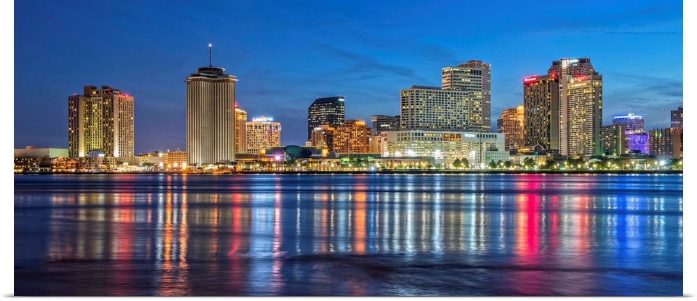 Photograph of the New Orleans skyline lit up at dusk and reflecting colorful bands onto the Mississippi River.