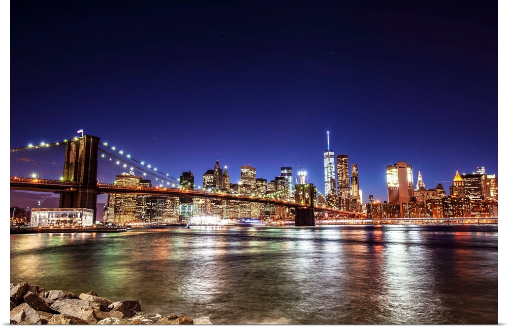 View of the New York City skyline illuminated at night, with the Brooklyn Bridge, from across the water.