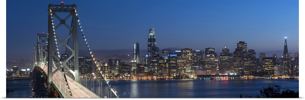 Panoramic photograph of the Bay Bridge and the San Francisco skyline lit up at dusk.
