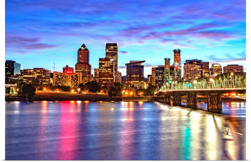 Photograph of the Portland, Oregon skyline lit up at sunset with colors reflecting onto the Willamette River in the foregr...