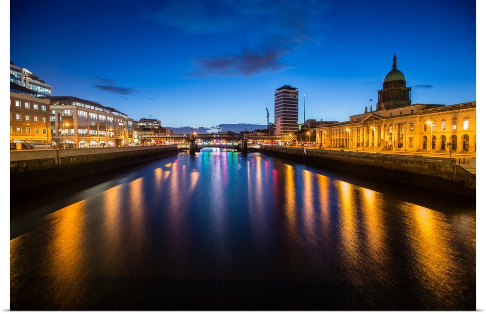 Photograph going straight down the River Liffey with buildings, highlighting The Custom House, on the sides lit up at night.