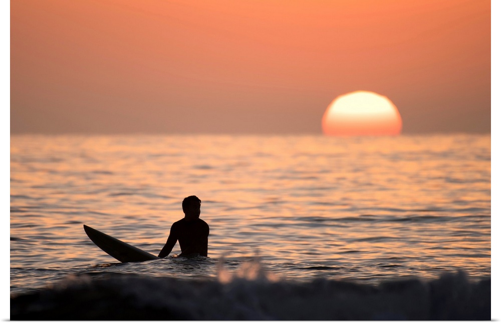 Silhouetted photograph of a man sitting on his surfboard in the Pacific Ocean on the coast of San Diego, California with a...