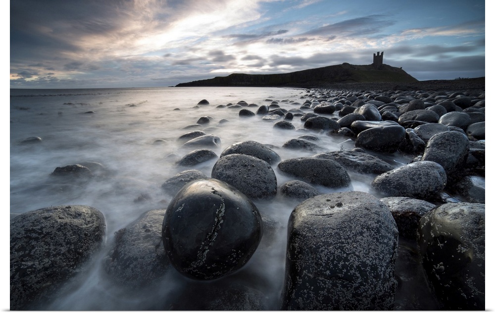 View at sunrise towards the ruin of Dunstanburgh Castle from the beach of basalt boulders known as The Rumble Churn at the...