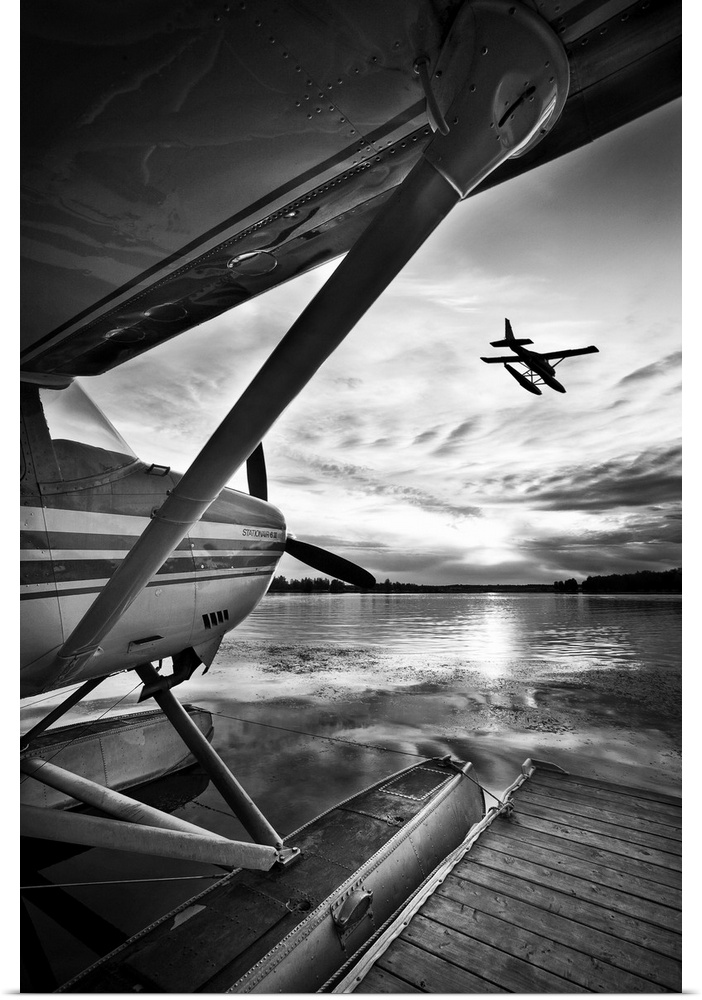 A black and white photograph taken from under the wing of a plane docked in the water while another plane has just taken o...