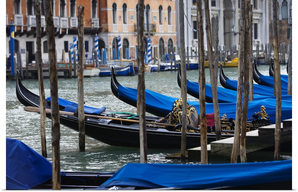 Photograph taken of a river in Venice that is lined with gondola boats that are tied up and have been covered with blue cl...