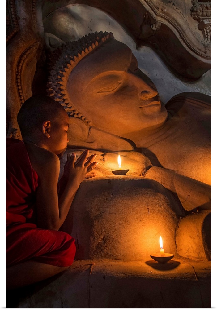 Young Burmese monk praying by candlelight by large reclining Buddha.