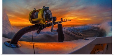 The reel of a big game fishing rod on the side of a boat, with the setting sun behind
