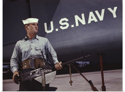 Sailor holding a .30-caliber machine gun in front of a U.S. Navy plane at Naval Air Base