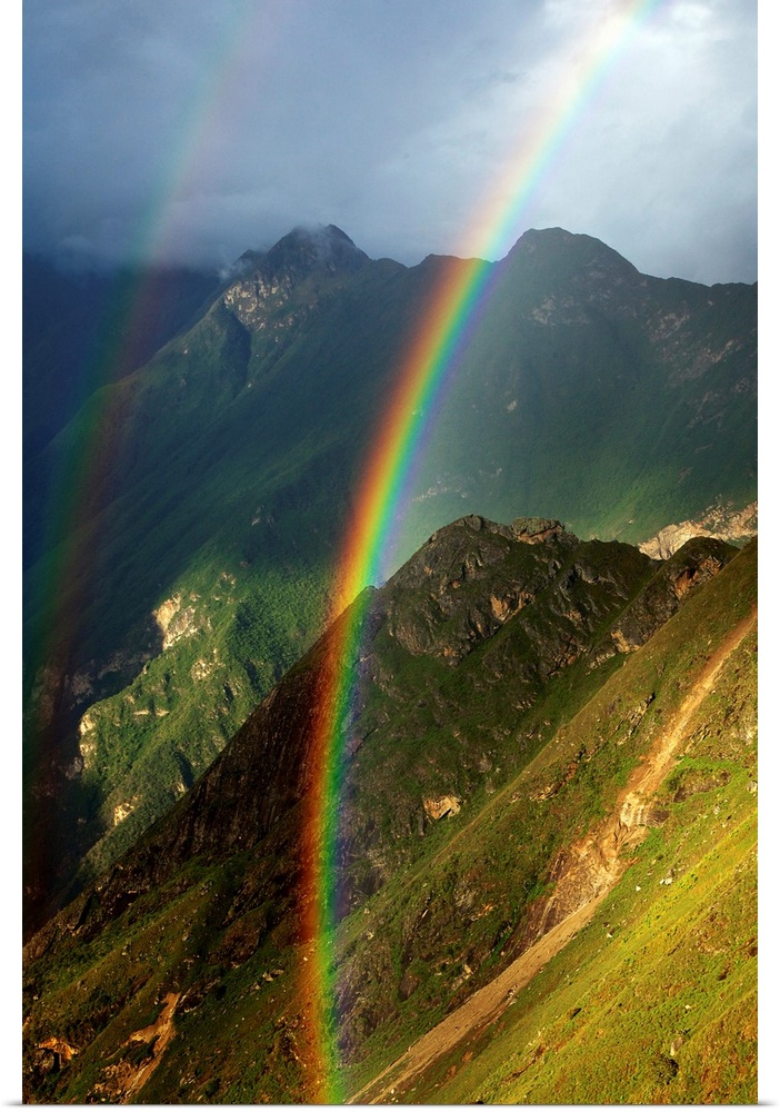 Vertical photograph of a double rainbow over green mountains on a cloudy day.