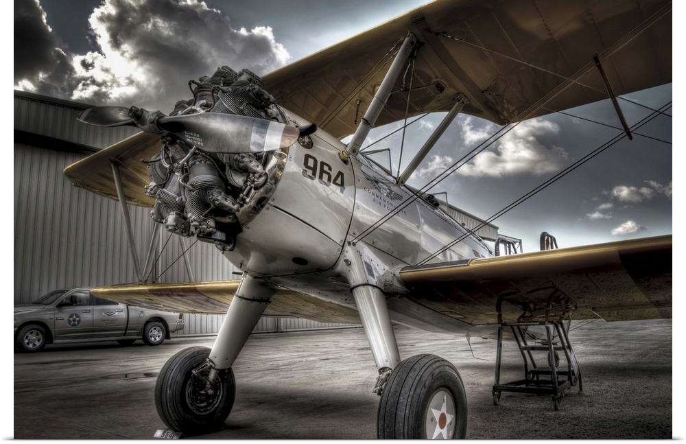 A HDR photograph of a vintage air craft parked on the tarmac outside a hanger and a sky full of large cumulous clouds. The...