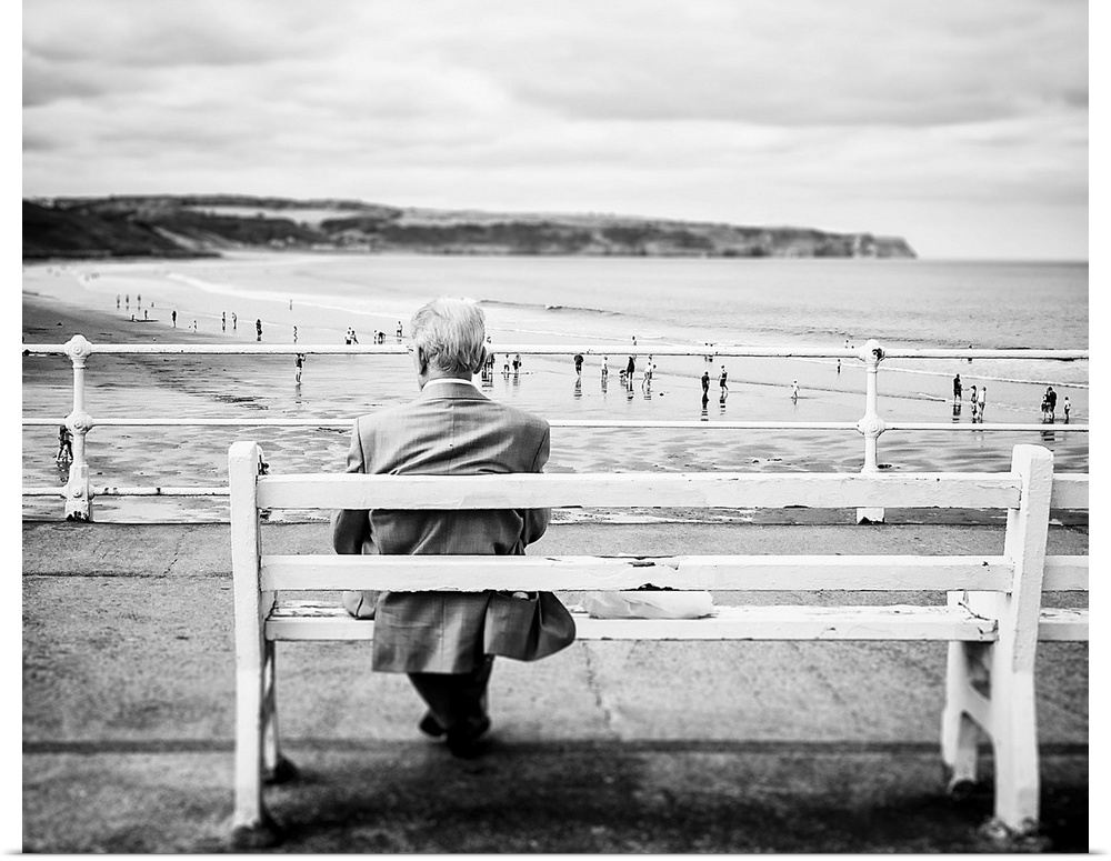 An old man sits gazing out to sea, on a white bench at a seaside resort.  Families can be seen on the beach beyond with cl...