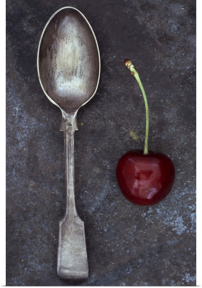 Antique tarnished silver teaspoon lying next to single dark red cherry with stalk on tarnished metal