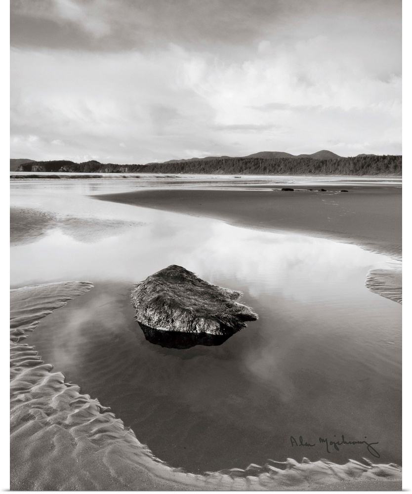 A black and white photograph of an idyllic beach scene, with a large rock in the foreground.