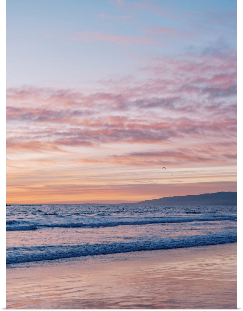 A photograph of gentle waves lapping the beach under an early evening sky where the sun is beginning to set.