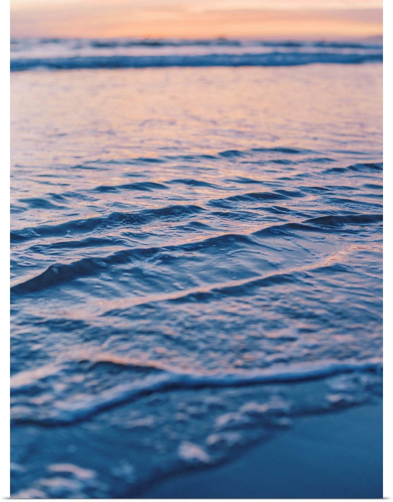 A close up photograph of gentle waves lapping the beach under an early evening sky where the sun is beginning to set.