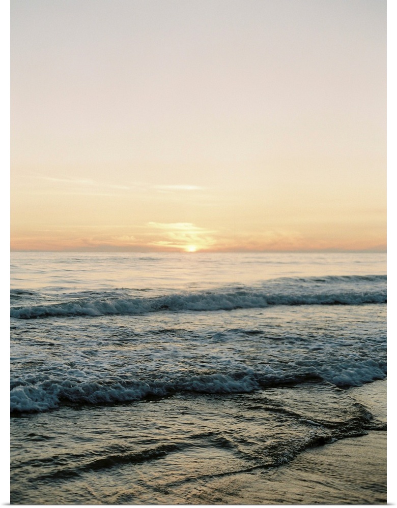 A photograph of gentle waves lapping the beach under an early evening sky where the sun is beginning to set.