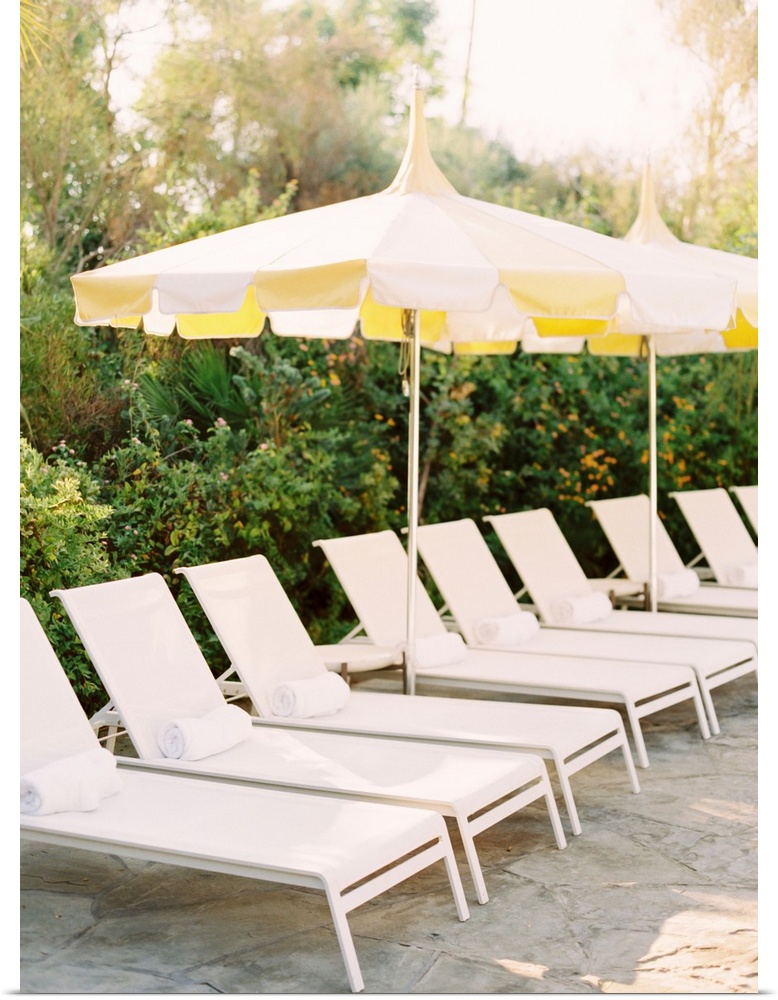 A photograph of a neat row of pool loungers with rolled towels underneath yellow umbrellas.