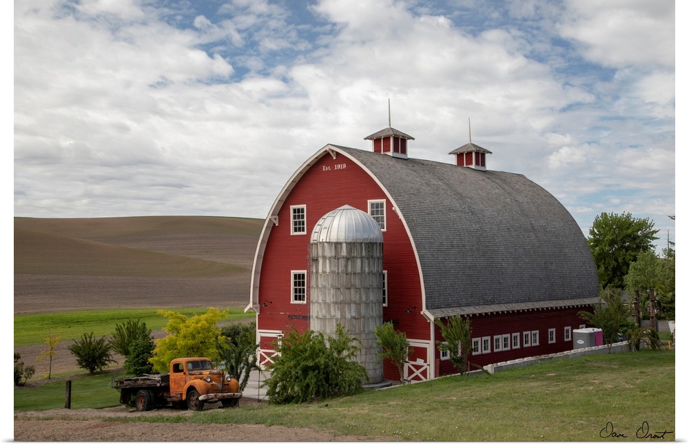 A photograph of a large red barn and grain silo with a vintage orange truck parked out front