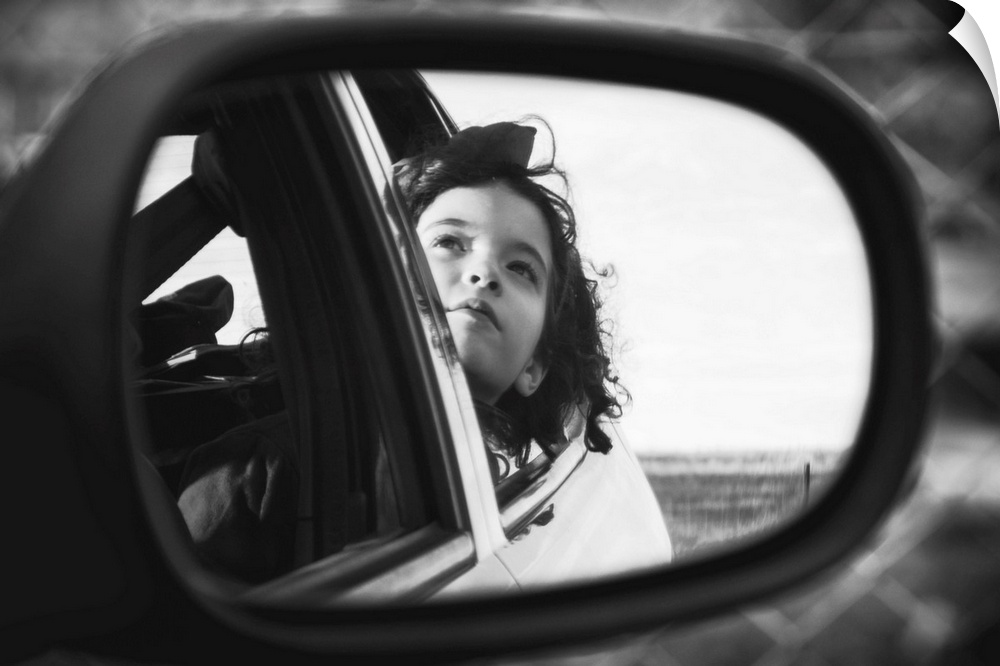 Black and white photo of a young child looking out a car window, seen from a rearview mirror.