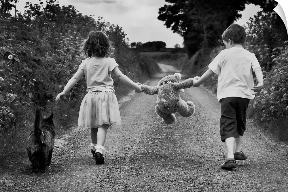 A young boy and girl each holding the arm of a teddy bear walk with their dog along a path.