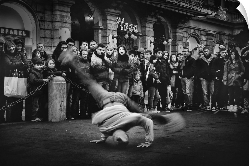 A breakdancer spins on their head while performing in the street, while onlookers watch.