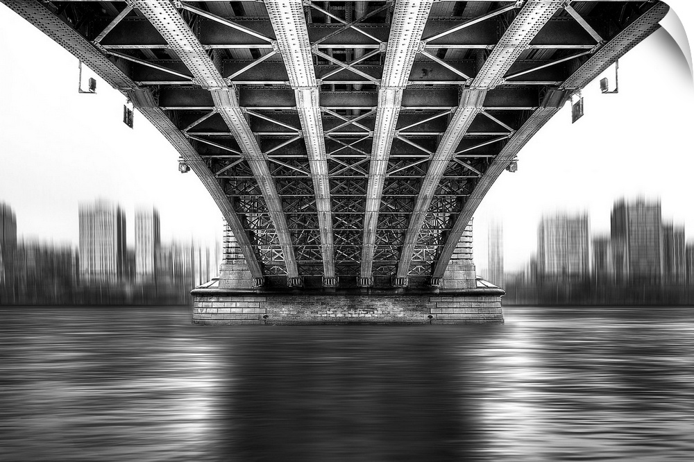 A black and white photograph from under a large bridge looking across the river it spans to see a city skyline.