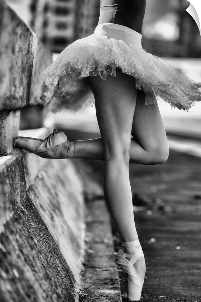 A black and white photograph of a ballerina in a dancers pose leaning against a road divider outdoors.