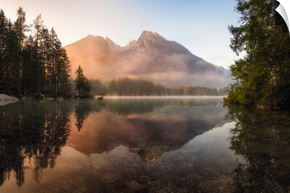 Reflective landscape photograph on a lake with trees and mountains in the background at sunrise.