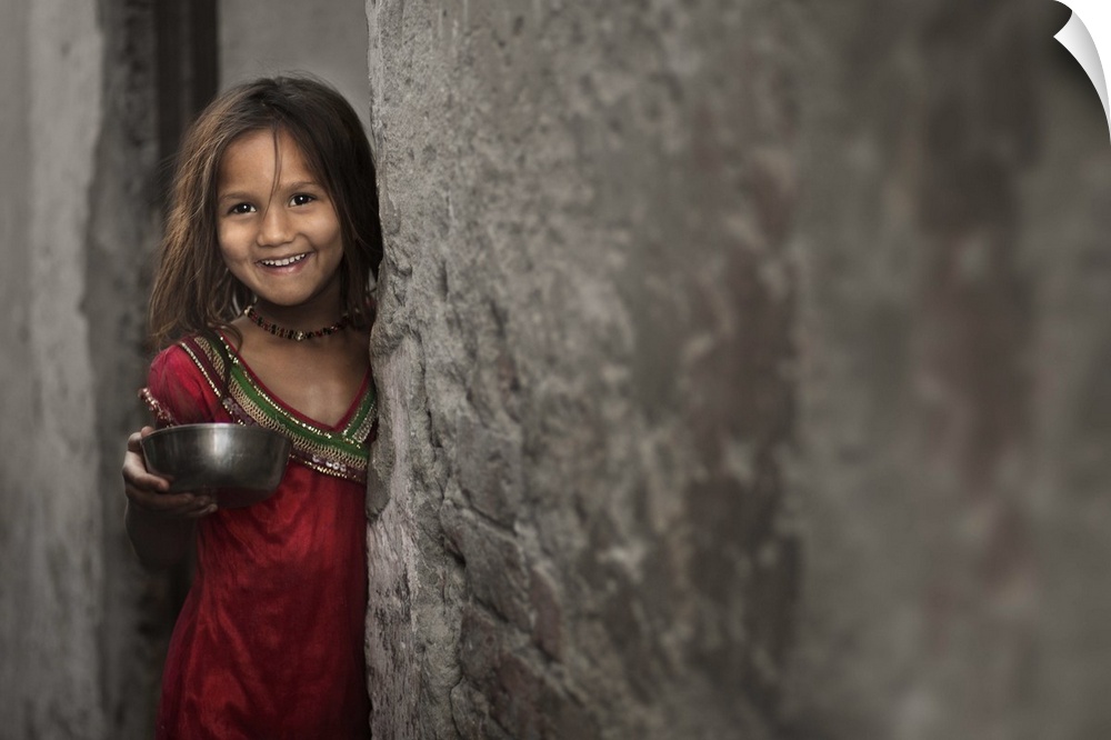 A smiling little girl holding a bowl, standing beside a stone wall.