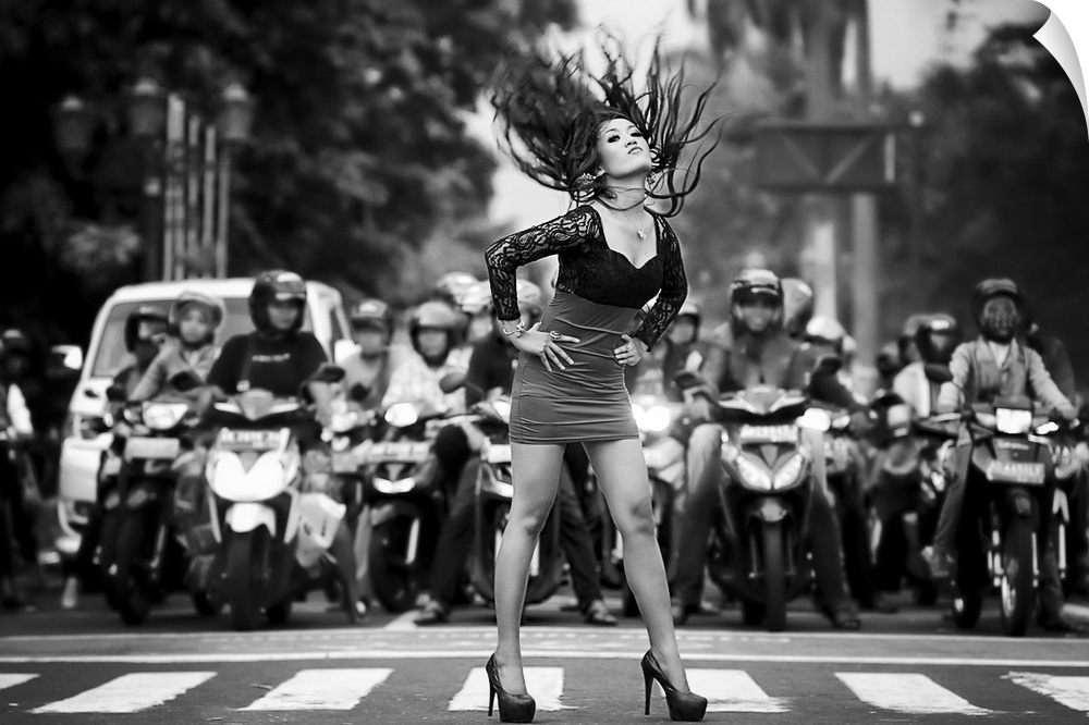 A woman in heels and a dress throws her hair back in a street crossing, with several motorcyclists behind her.