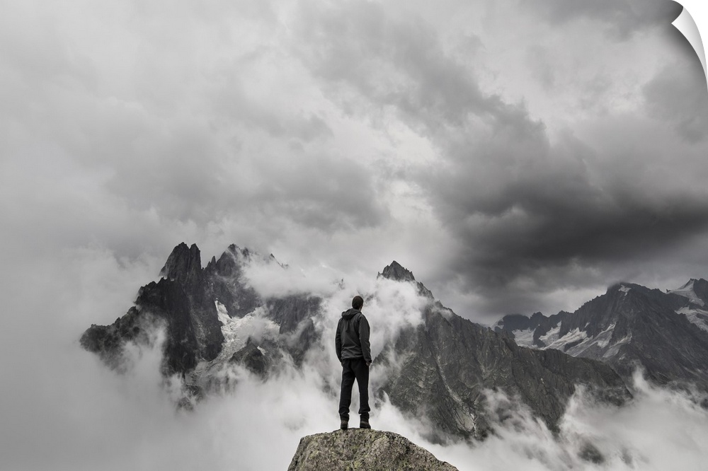A portrait of a man standing on a large rock formation staring off into the distance at a cloudy mountain range.