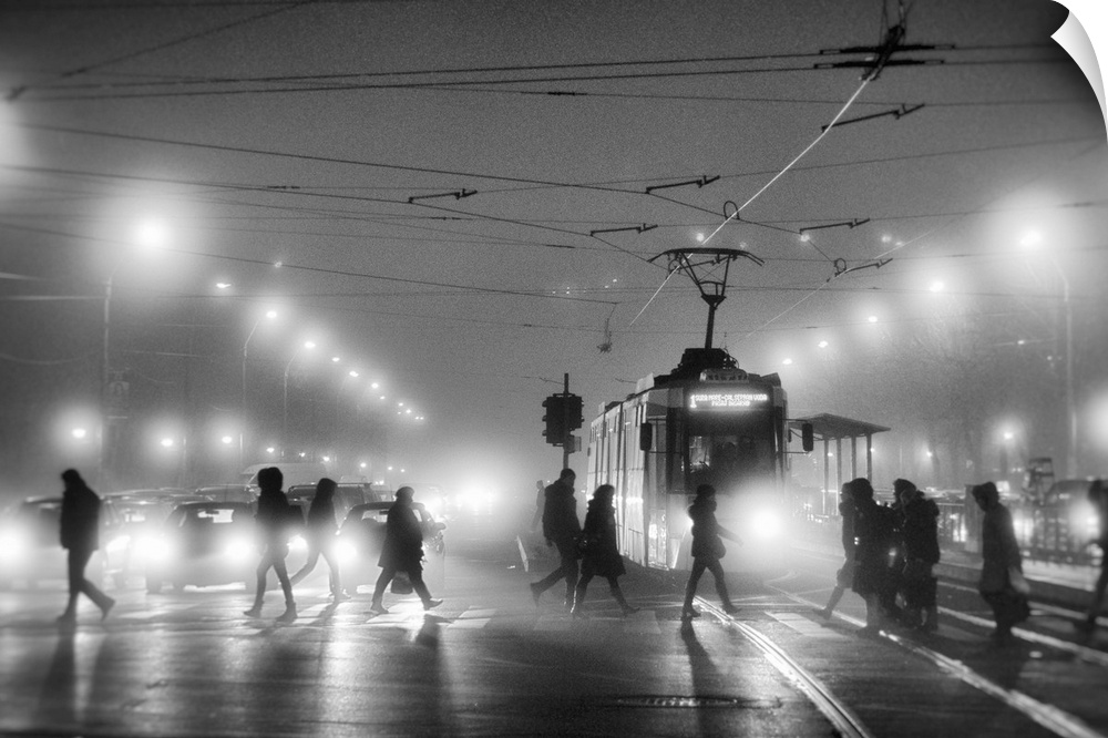 A group of people cross the street in a city at night, while cars and trams wait to pass.