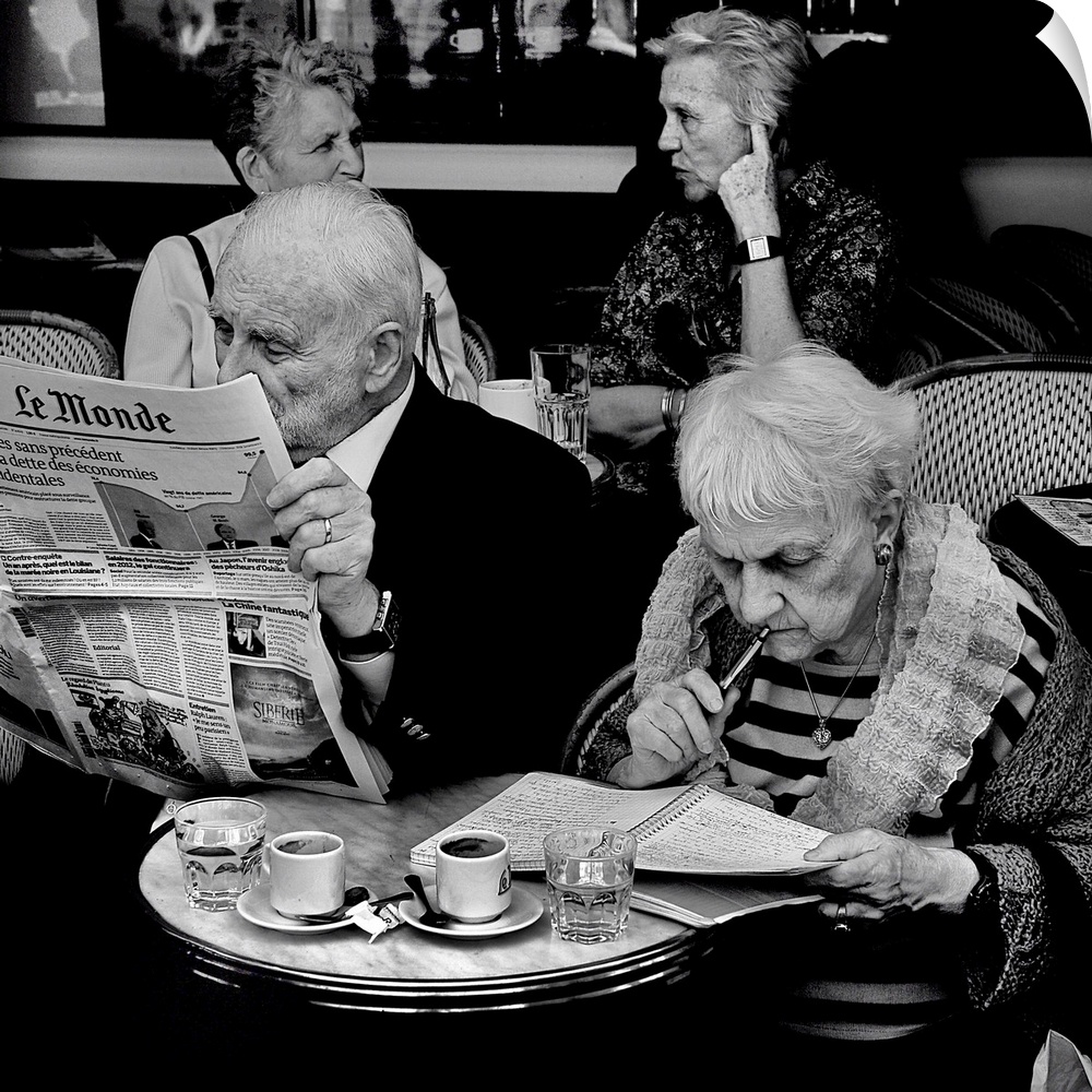 An elderly couple sit at a cafe. The man reads the morning news and the woman writes in a notebook.