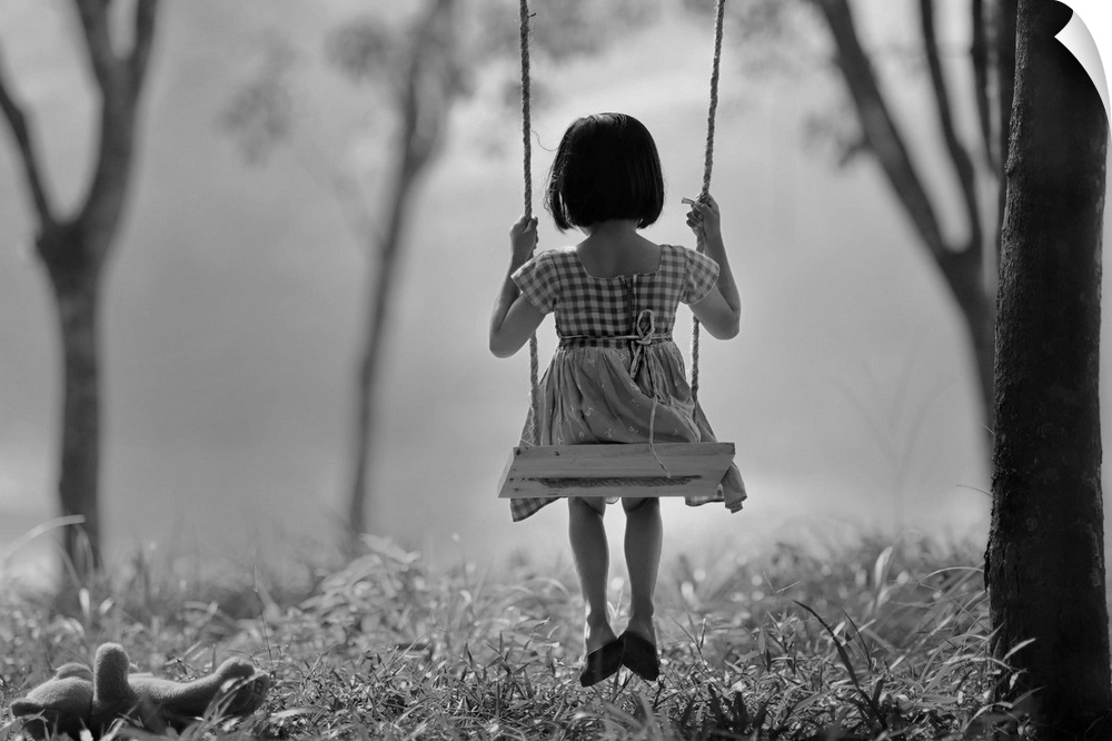 A young girl sitting on a swing in a forest, with a teddy bear on the ground.