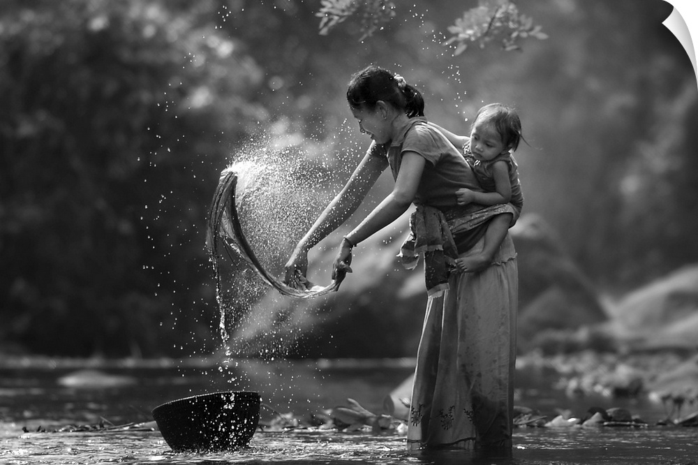 A mother in Indonesia with her child on her back washes clothing in a river.