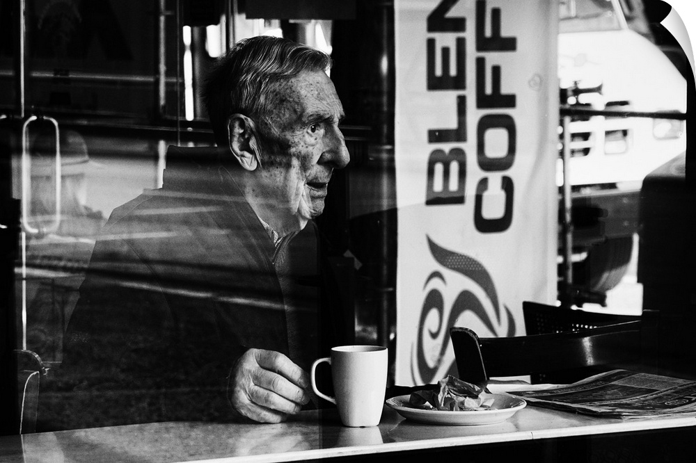 A photograph of an older man sitting in a cafe drink a cup of coffee.