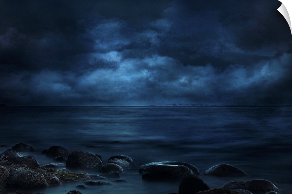 Blue landscape photograph of a rocky seashore lit by moonlight with a bridge in the far distance.