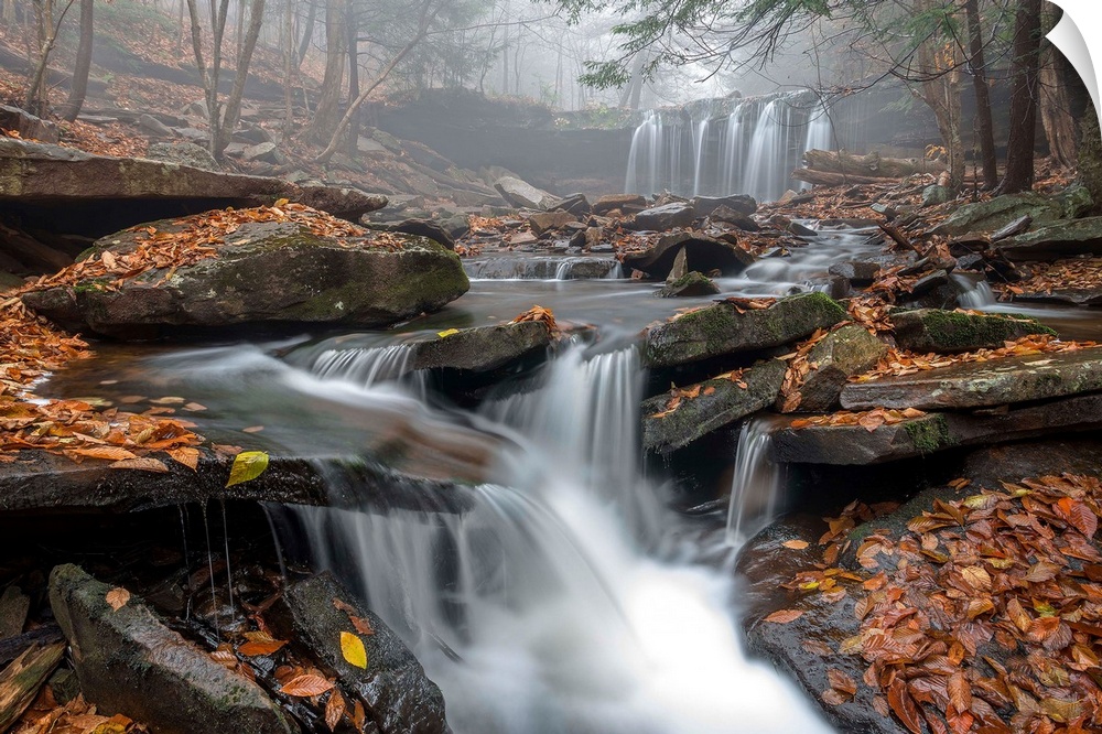 A waterfall and stream with a rocky bed and fallen leaves in a misty forest in Ricketts Glen, Pennsylvania.