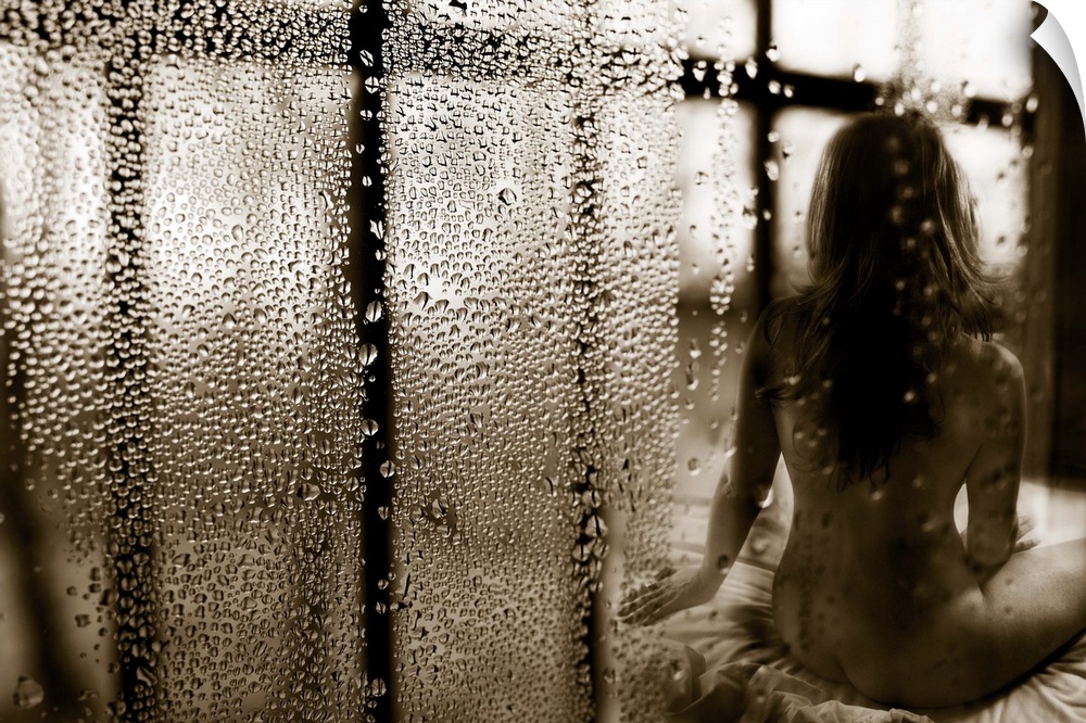 A nude woman sitting on a bed, with lots of raindrops on the window.