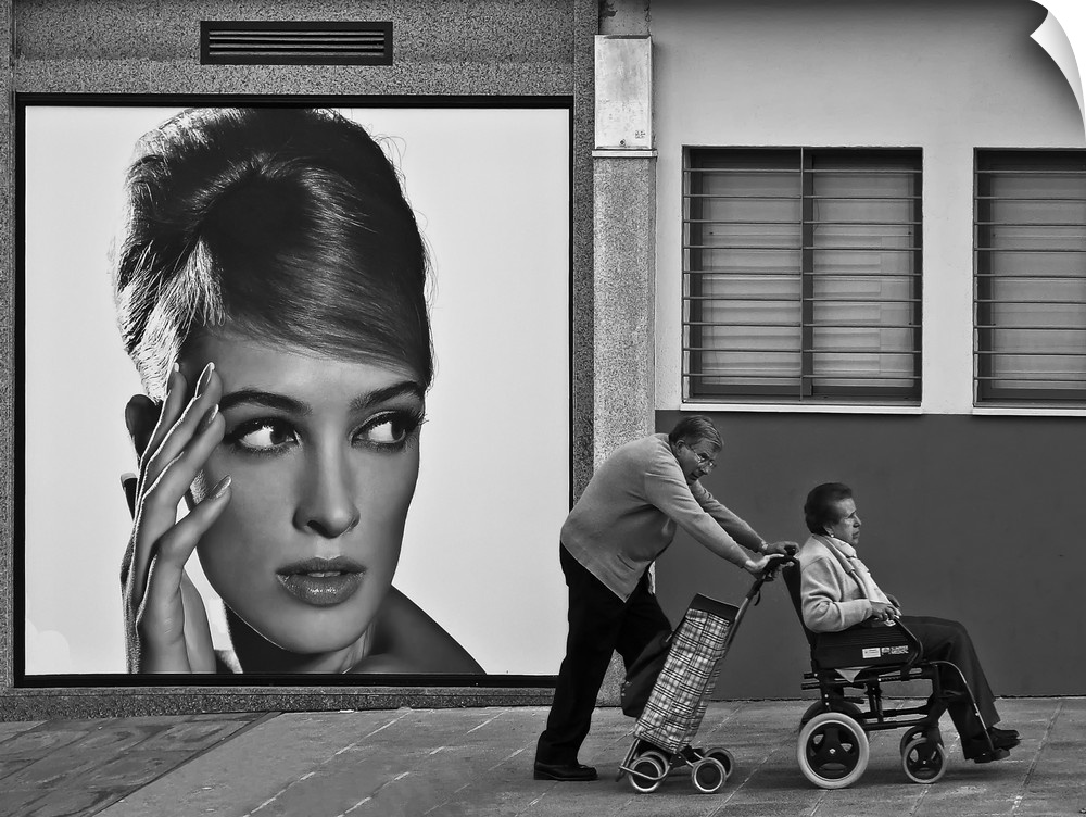 A man pushes his wife in her wheelchair along the street, past a large advertisement showing a young woman.