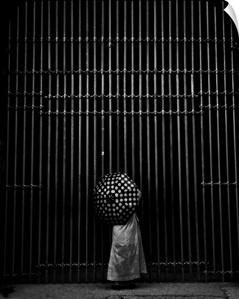 A person's polka-dotted umbrella contrasts with the vertical lines of an iron gate.