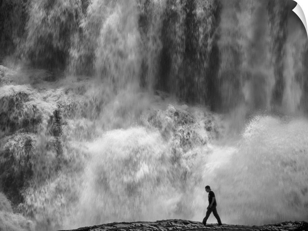 Man walking at the base of a huge water fall, looking tiny in comparison.
