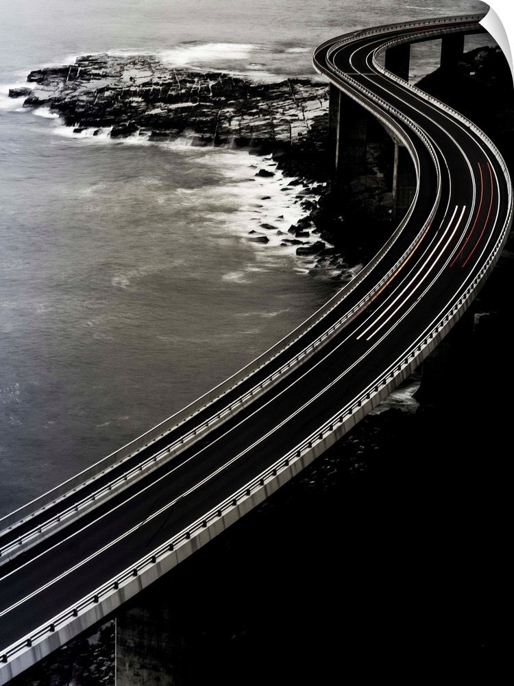 Vertical long exposure photograph of a winding bridge over an ocean with red and orange lines from traveling cars.