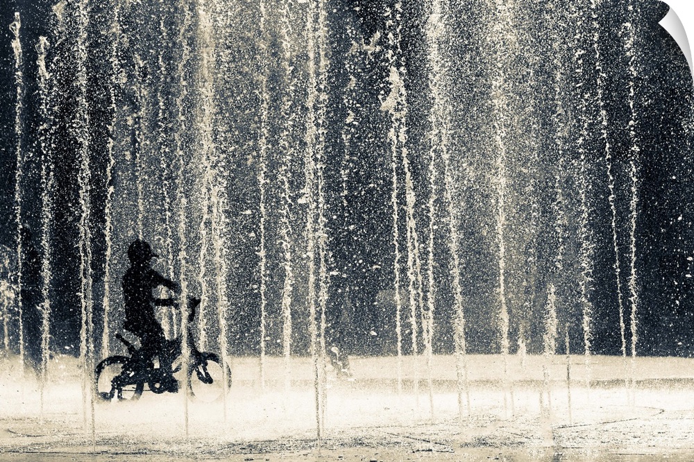 A child pedals his bicycle through numerous water jets of a fountain.