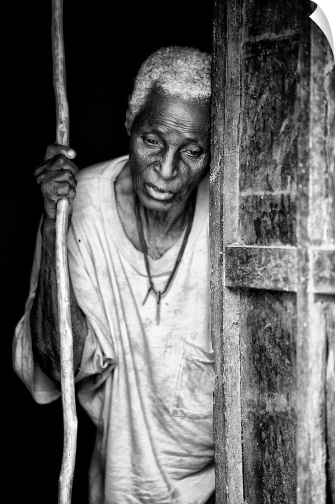 Black and white portrait of an elderly person with a staff in a doorway.