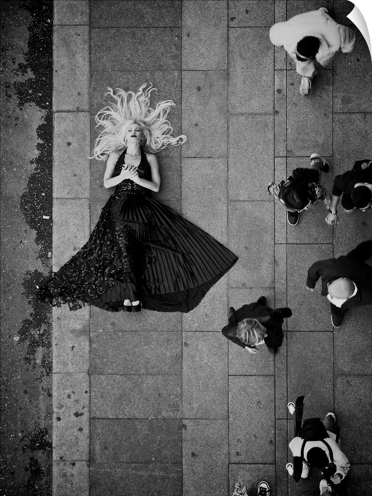 A beautiful blond woman in a flowing dress laying on the sidewalk as people walk by.